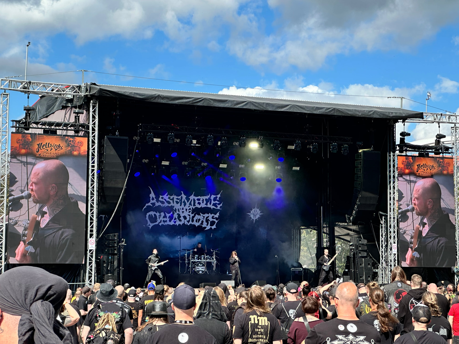 Assemble the Chariots performing on the outdoor stage at Hellsinki Metal Festival 2026 under a blue sky with their logo on the backdrop